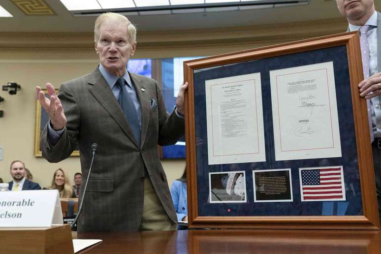 Bill Nelson, vestindo paletó e gravata, segura uma moldura contendo dois documentos, uma foto da superfície lunar e a bandeira americana.