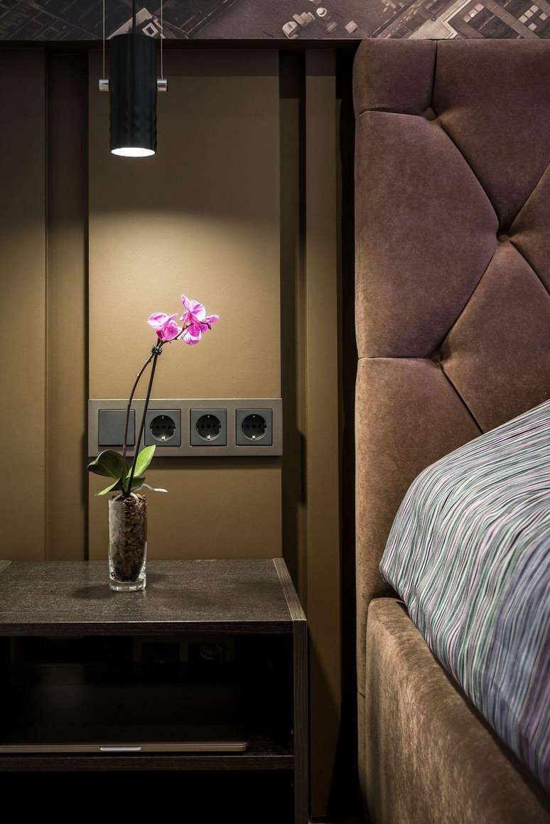 Detalhe de um quarto com parede na cor mocha mousse, cabeceira estofadabetana jogostom marrom, mesa de apoio com vaso de orquídea rosa, iluminação pendente e tomadas embutidas na parede