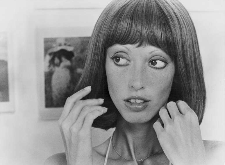 Shelley Duvall em retrato branco e preto, mãos levantadas, com quadro ao fundo