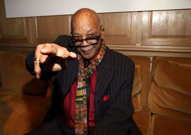 Quincy Jones usa óculos e aponta para câmera em retrato. Está sentado em um sofá/