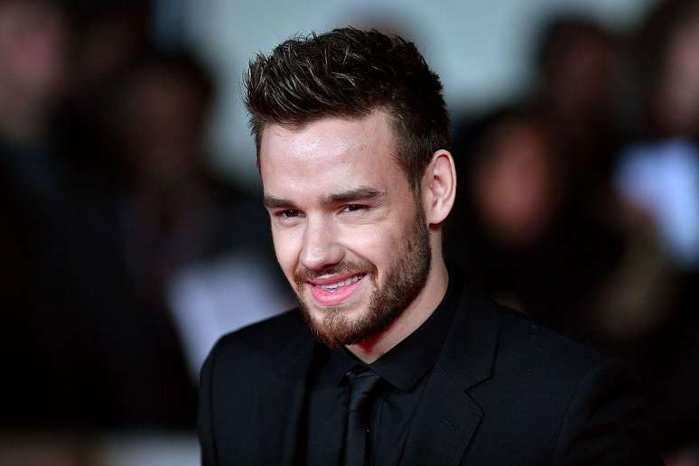 Liam James Payne veste roupa social e sorri para imagem com fundo desfocado