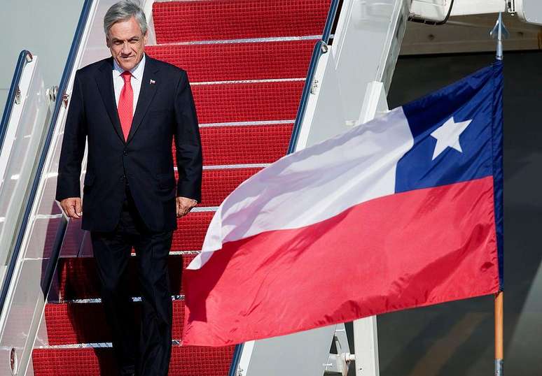 A imagem mostra Sebastián Piñera, ex-presidente chileno, descendo as escadas de um avião, com a bandeira do Chile ao lado, simbolizando sua liderança no país durante dois mandatos.