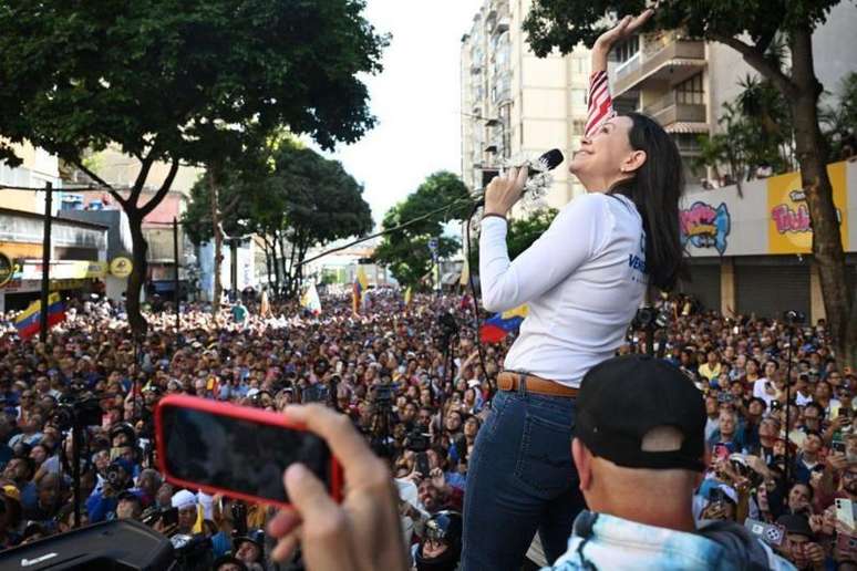 Corina discursando no microfone e sorrindo ao olhar pra cima; ela é ouvida por centenas de pessoas na rua