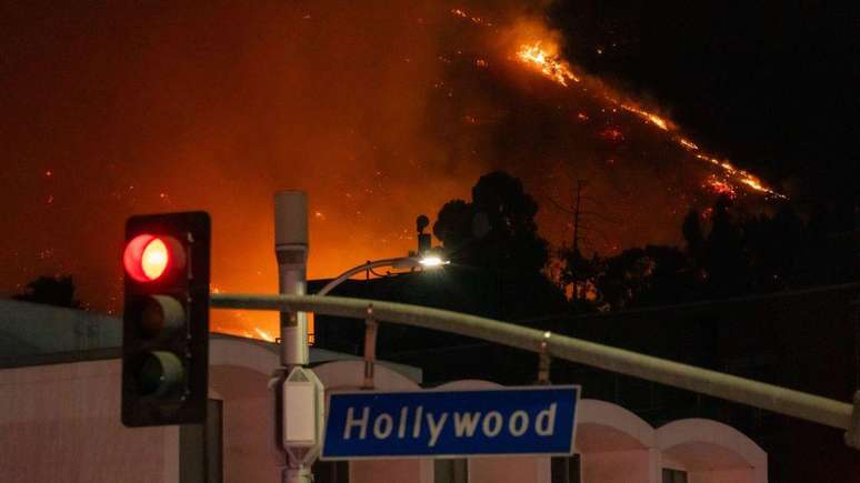 Placa de trânsito com localização de Hollywood em primeiro plano, com fogo ao fundo