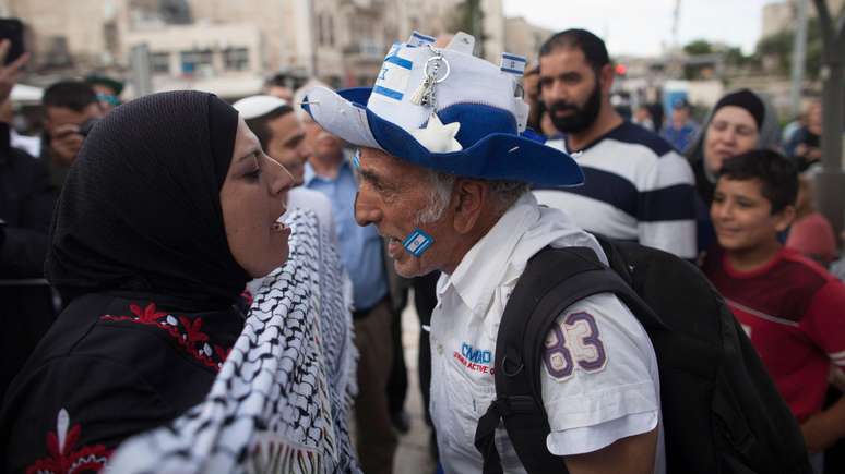 Uma mulher palestina usando um lenço na cabeça e um homem israelense usando um chapéu azul e branco com a bandeira israelense gritam um com o outro em Jerusalém