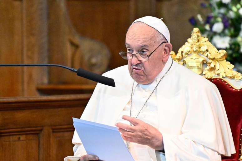 O Papa Francisco sentado em uma cadeira de veludo, lendo de uma folha de papel. Ele está falando em um microfone. O fundo está desfocado