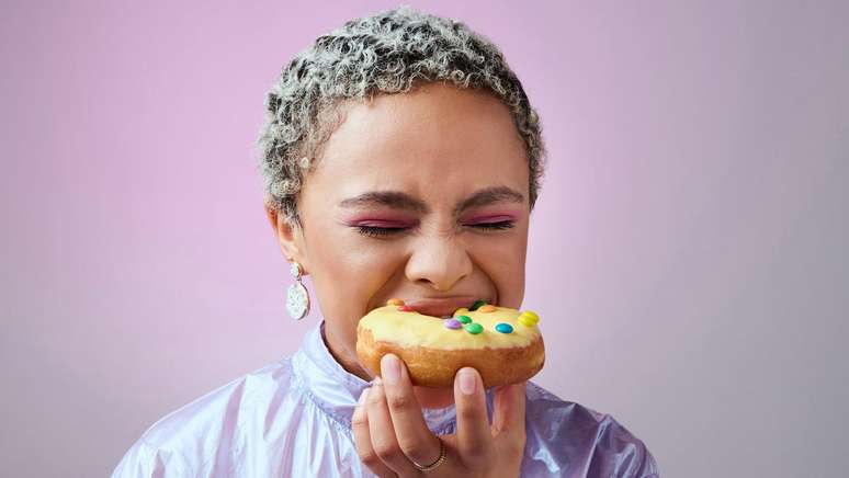 Mulher negra, de cabelos curtos e claros, mordendo um donut com cobertura amarela