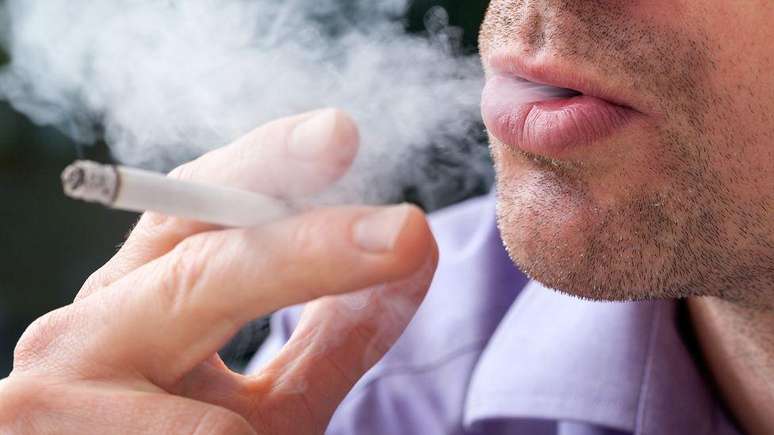 Foto de um homem fumando um cigarro. Ele tem um cigarro na mão e está soprando fumaça para fora da boca.