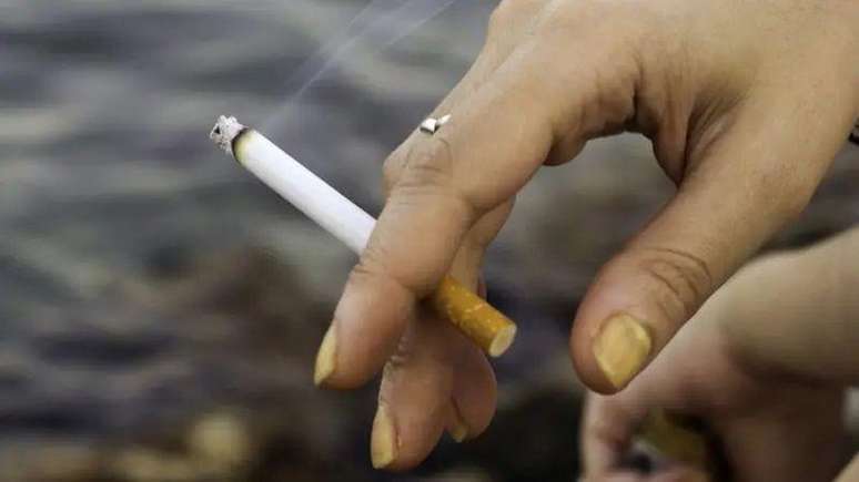 Foto de uma mão com unhas amareladas segurando um cigarro. 