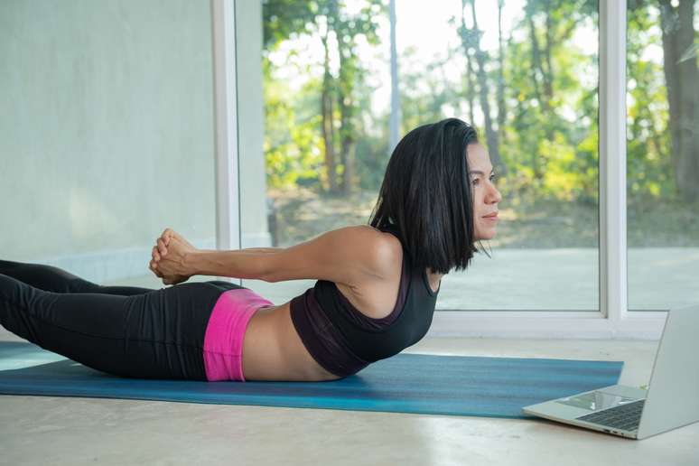Para evitar descompensações musculares que acarretam dores na lombar e ciático, é preciso fortalecer os músculos do abdome, quadril e pélvisstretching watching fitness video tutorial online on laptop, doing workout at home sitting on mat in living room practicing