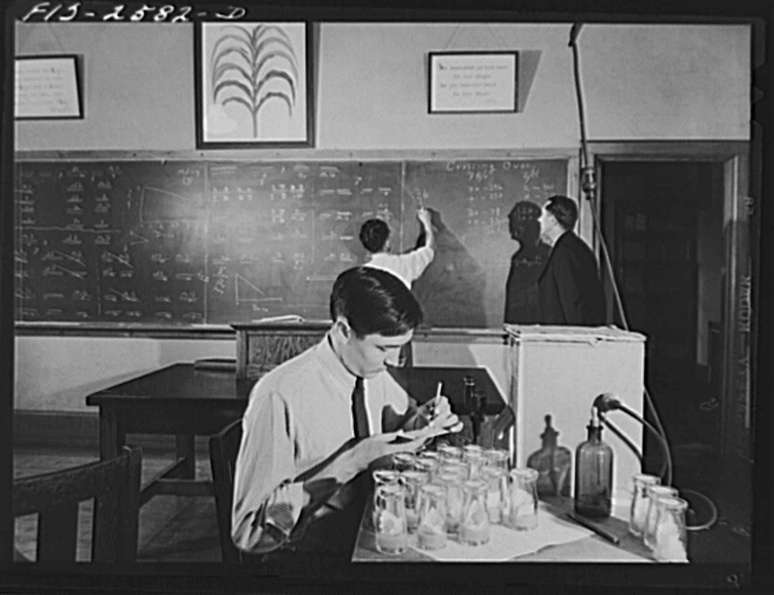 Homem sentado de camisa e gravata faz anota&ccedil;&otilde;es acima de jarros, dois outros homens em um quadro-negro com um p&ocirc;ster de milho acima