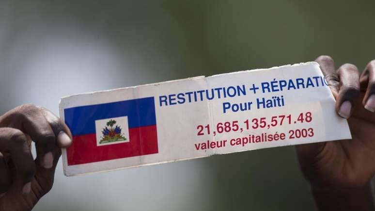 Manifestante segura um cartaz em francês com os dizeres “Restituição e compensação para o Haiti” enquanto ouve o discurso do presidente francês François Hollande do lado de fora do Palácio Nacional em Porto Príncipe, Haiti, em maio de 2015. (Imagem ilustrativa)