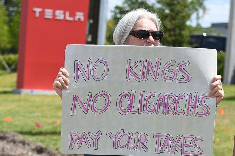 Uma mulher segura um cartaz em frente a uma concessionária da Tesla com os dizeres: "Sem reis, sem oligarcas — paguem seus impostos"