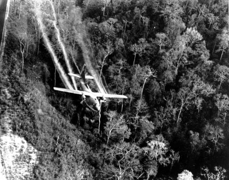 Um avião sobrevoa a selva, com jatos de desfolhante fluindo atrás dele.