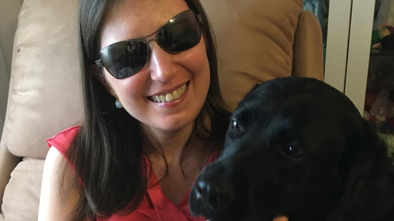 Danieli Haloten sentada com a labradora preta Elva, sua cão-guia aposentada, em seu colo em uma foto publicada no Instagram