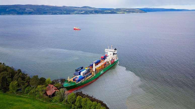 Vista aérea do navio verde, cheio de contêineres, com o topo do barco em terra, ao lado da casa.