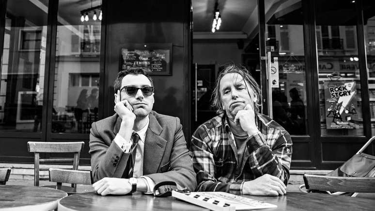 Dois homens com o bra&ccedil;o apoiado sobre uma mesa ao ar livre em foto em preto e branco