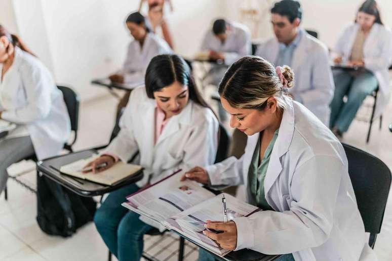 Estudantes de medicina sentados em carteiras, vestindo jalecos brancos, discutindo um livro did&aacute;tico.