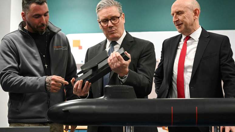 O primeiro-ministro britânico Keir Starmer, ao centro, e o secretário de Defesa John Healey, à direita, se reúnem com aprendizes do sistema BAE enquanto observam um modelo de submarino durante uma visita conjunta em Barrow-in-Furness, Inglaterra, na quinta-feira, 20 de março de 2025.