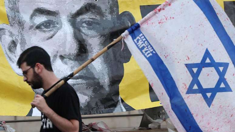 Um homem com uma bandeira israelense passa por um cartaz com a foto de Netanyahu