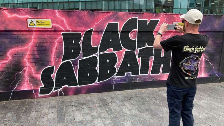 Um f&atilde; com uma camiseta do Black Sabbath tirando uma foto de um grande mural colorido do Black Sabbath em uma parede em Birmingham