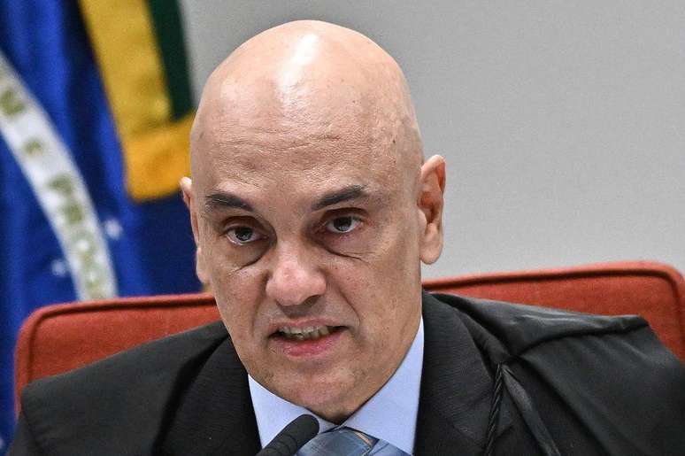 Alexandre de Moraes falando ao microfone em audi&ecirc;ncia, com bandeira do Brasil atr&aacute;s