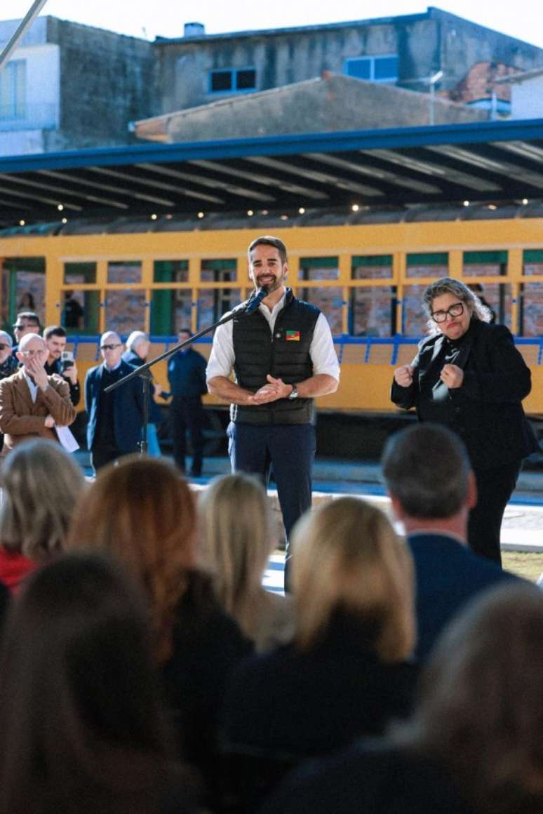 A foto mostra o governador Eduardo Leite discursando em um microfone em um palco ao ar livre. Ele est&aacute; sorrindo, de colete preto e camisa branca. H&aacute; uma mulher loira, como int&eacute;rprete, ao seu lado. Ao fundo, um pr&eacute;dio antigo de cor amarela, com janelas grandes, pode ser visto.