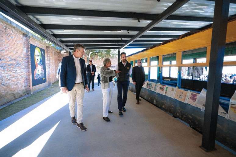 A foto mostra o governador Eduardo Leite (de casaco azul-escuro e cal&ccedil;a clara) caminhando ao ar livre por uma passarela coberta. Ele est&aacute; em grupo, e ao lado da passarela, h&aacute; um pr&eacute;dio antigo de cor amarela com janelas grandes. Na parede de tijolos ao lado, h&aacute; uma pintura mural com o rosto da Monalisa.