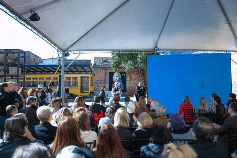 A foto mostra um evento sendo realizado ao ar livre. Em primeiro plano, &eacute; poss&iacute;vel ver a plateia sentada em cadeiras pretas sob uma grande tenda branca. No palco, h&aacute; um tel&atilde;o azul, e ao fundo, &agrave; esquerda, pode ser vista uma estrutura amarela e uma parede de tijolos com uma pintura mural, uma releitura da Mona Lisa. No palco, duas mulheres est&atilde;o em p&eacute;, e uma delas, de preto, est&aacute; falando ao microfone.