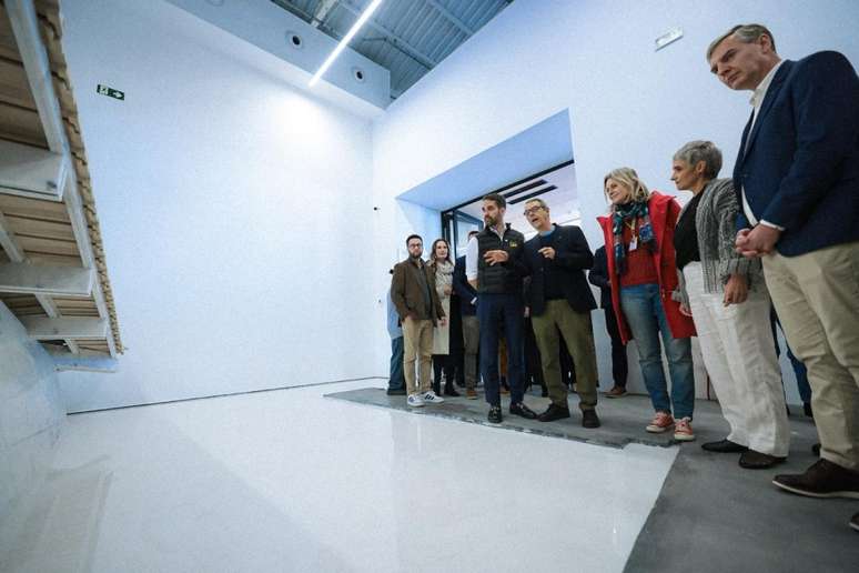 A foto mostra o governador Eduardo Leite e outras pessoas em uma sala com paredes brancas e piso branco. O grupo est&aacute; olhando para uma escultura de madeira que se estende por uma das paredes. O governador est&aacute; de lado, olhando para a obra de arte.