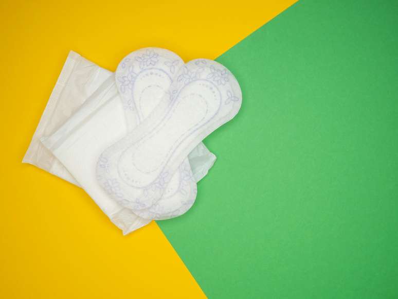 A imagem mostra absorventes brancos sobre um fundo amarelo e verde.