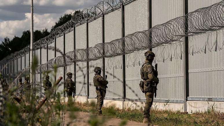 Soldados de fronteira armados montam guarda frente a uma alta e longa barreira metálica com concertinas de arame farpado