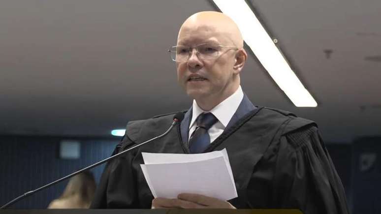 Demóstenes segurando papéis e falando ao microfone em sala de audiência