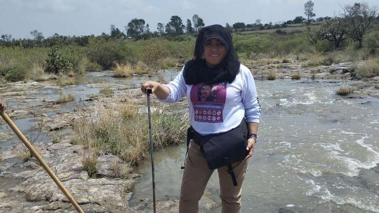 Yadira Gonz&aacute;lez, de p&eacute;, perto de um riacho, vestindo uma camisa com a imagem do seu irm&atilde;o. Ela sorri para a c&acirc;mara e segura uma esp&eacute;cie de bast&atilde;o, como os usados para caminhar nas montanhas.