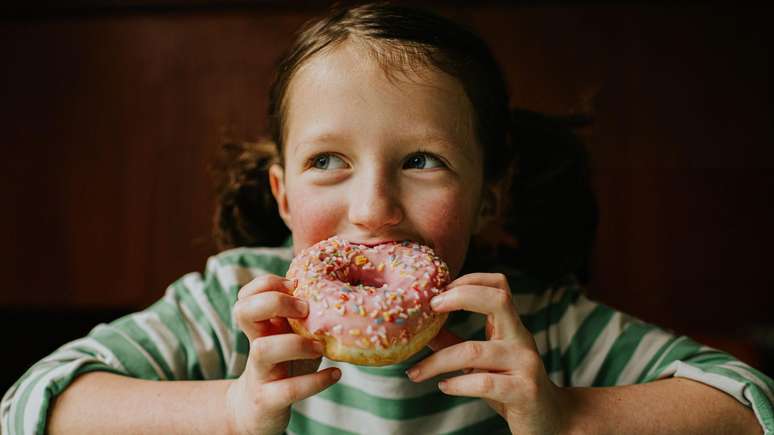 Crian&ccedil;a com cabelo castanho, vestindo uma camiseta listrada verde e branca, morde um donut com creme rosa e enfeites
