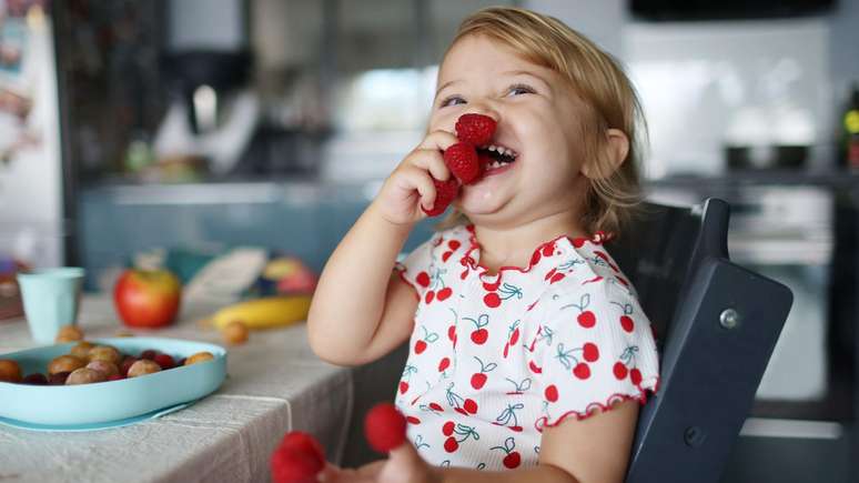 Crian&ccedil;a com cabelo loiro, vestindo uma camiseta com estampa de cerejas, ri enquanto segura framboesas enfiadas nos dedos das duas m&atilde;os