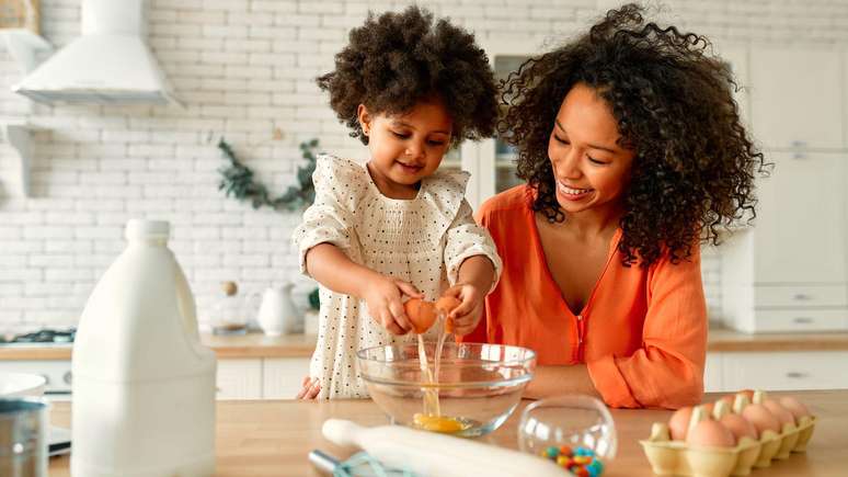 Crian&ccedil;a e sua m&atilde;e, sentadas &agrave; mesa da cozinha. A m&atilde;e veste uma blusa laranja brilhante e a crian&ccedil;a usa um vestido branco de bolinhas, enquanto quebra um ovo em uma tigela.