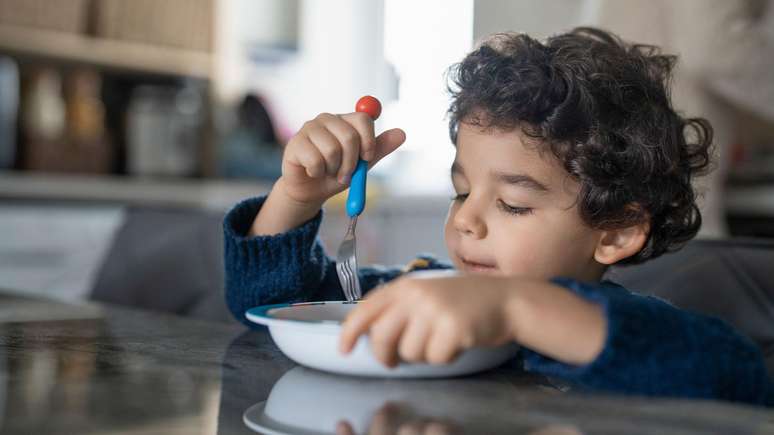 Crian&ccedil;a com cabelo ondulado marrom, vestindo uma blusa azul escura, comendo com um garfo em uma tigela branca