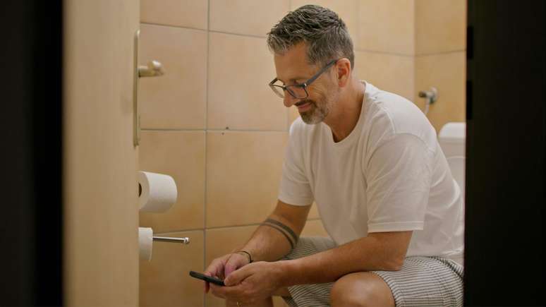 Homem sentado no vaso sanit&aacute;rio com seu telefone celular