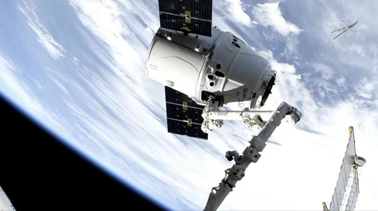 Uma nave espacial c&ocirc;nica branca com dois pain&eacute;is solares retangulares no espa&ccedil;o, com a Terra ao fundo.
