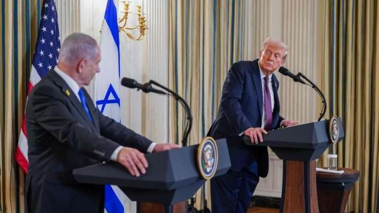 Netanyahu e Trump se olham, cada um de seu palanque em salão com bandeiras dos respectivos países