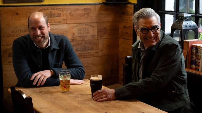 Pr&iacute;ncipe William e Eugene Levy sorriem enquanto bebem uma Guinness e uma cidra em um pub pr&oacute;ximo