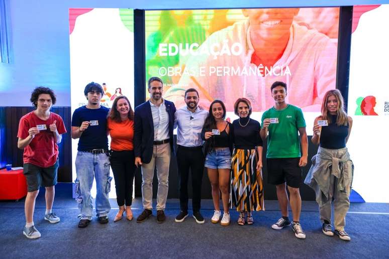 A imagem mostra um grupo de nove pessoas posando juntas em um palco, sorrindo para a foto. A maioria delas s&atilde;o jovens estudantes, segurando cart&otilde;es em m&atilde;os, como se tivessem acabado de receb&ecirc;-los. No centro do grupo, est&atilde;o tr&ecirc;s adultos &mdash; dois homens e uma mulher &mdash; que aparentam ser figuras p&uacute;blicas ou autoridades, provavelmente envolvidas na organiza&ccedil;&atilde;o do evento. O fundo do palco tem um painel eletr&ocirc;nico com uma imagem de um jovem sorridente e os dizeres "EDUCA&Ccedil;&Atilde;O - OBRAS E PERMAN&Ecirc;NCIA". &Agrave; esquerda e &agrave; direita do painel, aparecem logotipos coloridos que remetem ao Governo do Estado do Rio Grande do Sul, sugerindo que se trata de um evento oficial ligado &agrave; educa&ccedil;&atilde;o p&uacute;blica. As pessoas est&atilde;o vestidas de maneira casual, exceto os dois homens ao centro que usam roupas mais formais. O ambiente &eacute; iluminado com luzes azuis, e o clima &eacute; de celebra&ccedil;&atilde;o ou lan&ccedil;amento de um novo programa educacional.