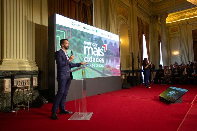 A imagem apresenta o governador falando ao microfone em frente a um grande painel digital com a frase “Avançar Mais Cidades” e logotipos de instituições como Cadip, Banrisul e BRDE.