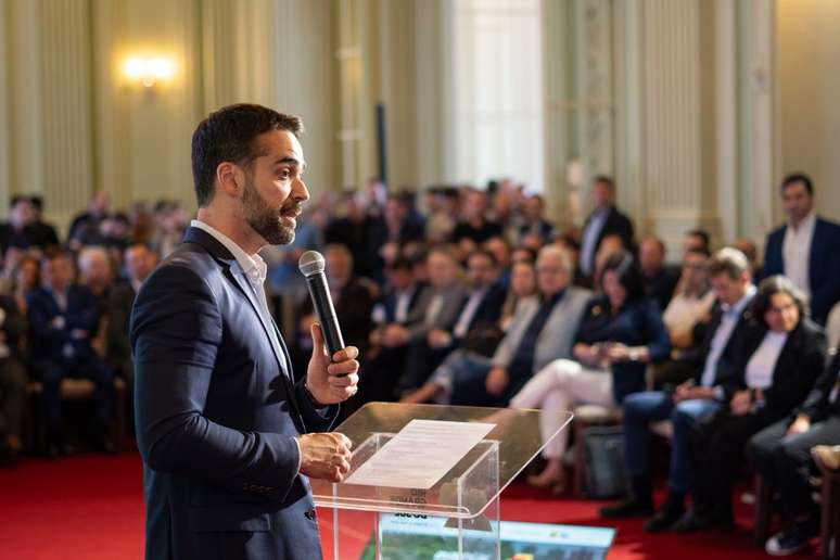 A cena mostra um homem em pé segurando um microfone, posicionado atrás de um púlpito transparente com papéis sobre ele. Ao fundo, há um público sentado em várias fileiras, todos vestidos formalmente.