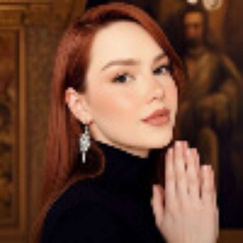 mulher ruiva de lado, com as m&atilde;os unidas pr&oacute;ximas ao rosto. ela veste blusa preta