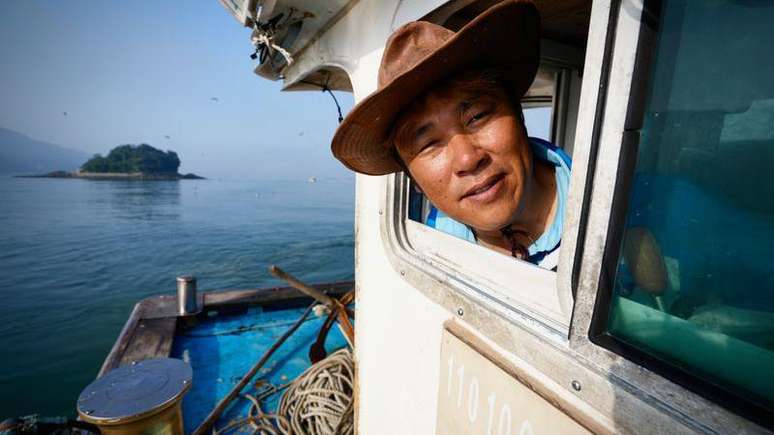 Um homem usando chap&eacute;u aparece com a cabe&ccedil;a para fora da janela de um barco. Ao fundo, v&ecirc;-se o mar e uma ilha.