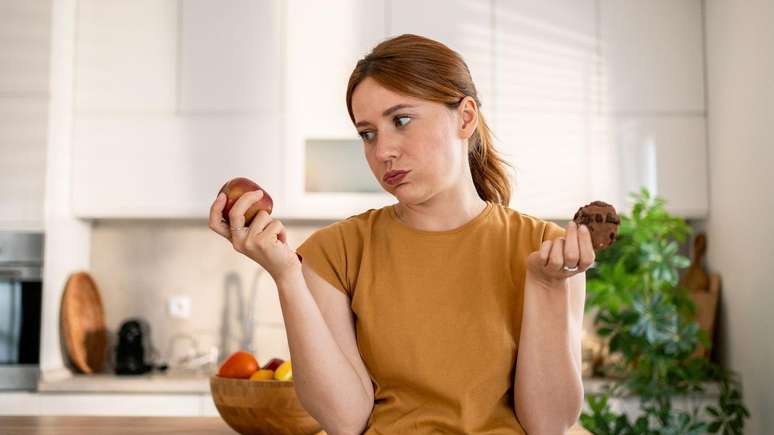 Young woman holding an apple and a chocolate chip cookie, contemplating a healthy food choice in a stylish modern kitchen. Young woman holding an apple and a chocolate chip cookie, contemplating a healthy food choice in a stylish modern kitchen.