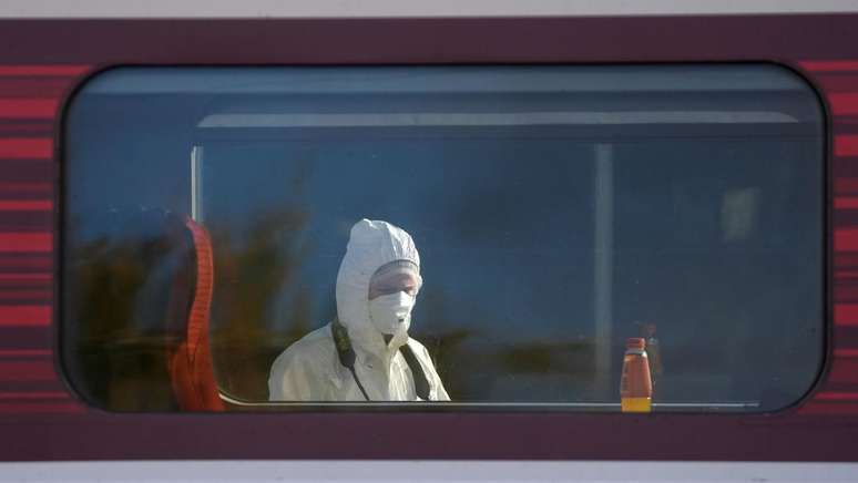 Cabe&ccedil;a e ombros de um perito policial s&atilde;o vistos atrav&eacute;s da janela de um vag&atilde;o de trem. Ele usa capuz branco sobre a cabe&ccedil;a e m&aacute;scara branca cobrindo nariz e boca.