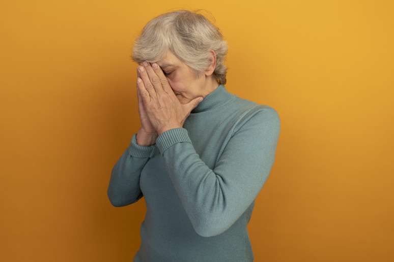 tired old woman wearing blue turtleneck sweater standing in profile view keeping hands together on face with closed eyes isolated on orange background with copy space